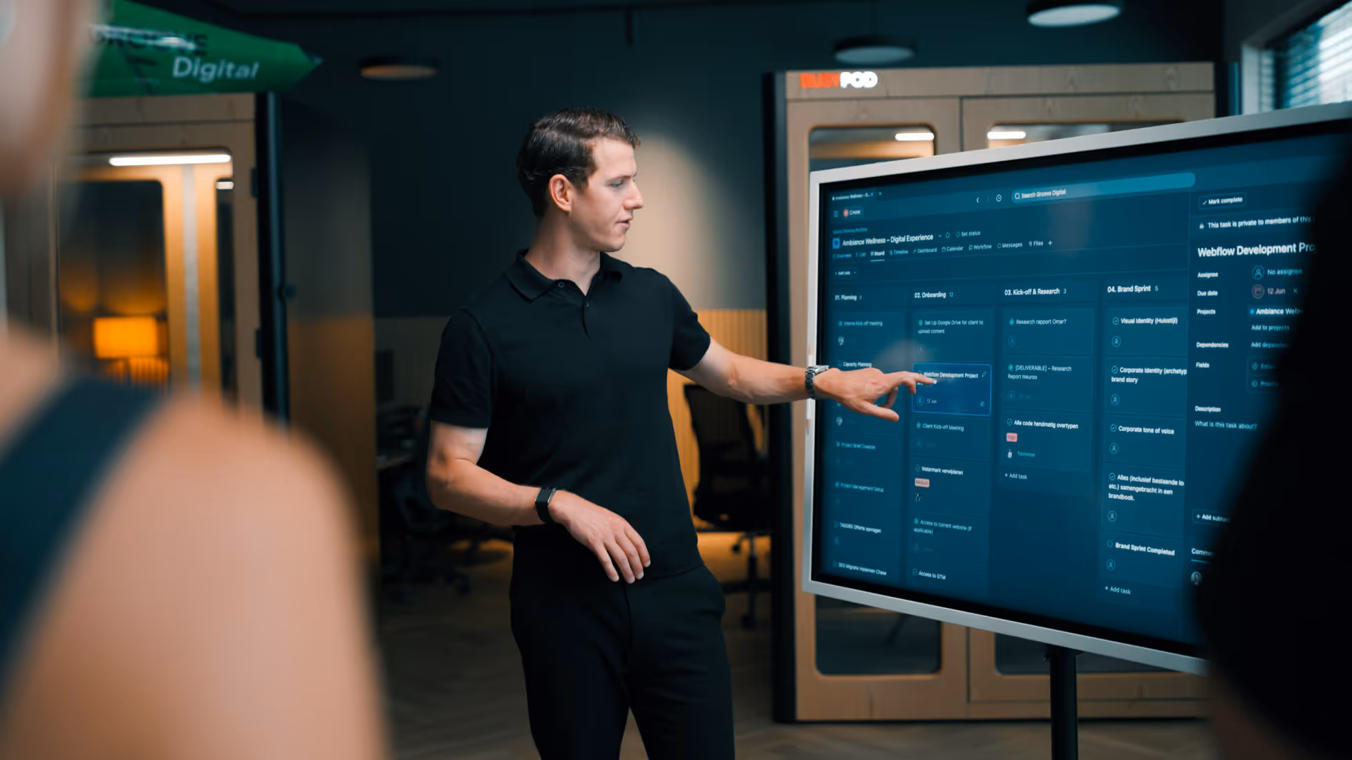 Man in zwart polo wijst naar informatie op groot touchscreen scherm in modern kantoor.