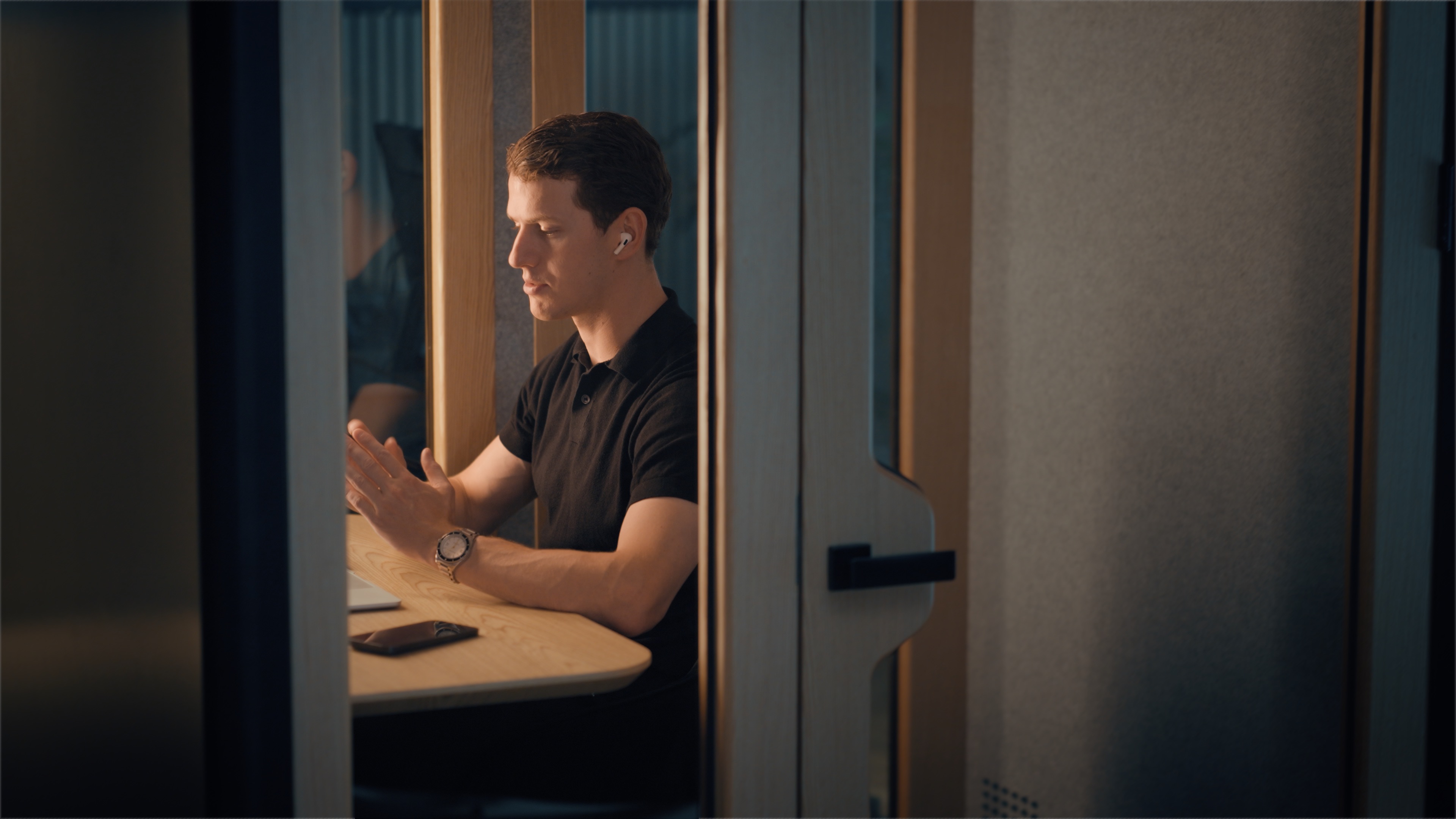Man met een zwarte polo gebruikt een laptop in een kleine, houten werkruimte met oordopjes.