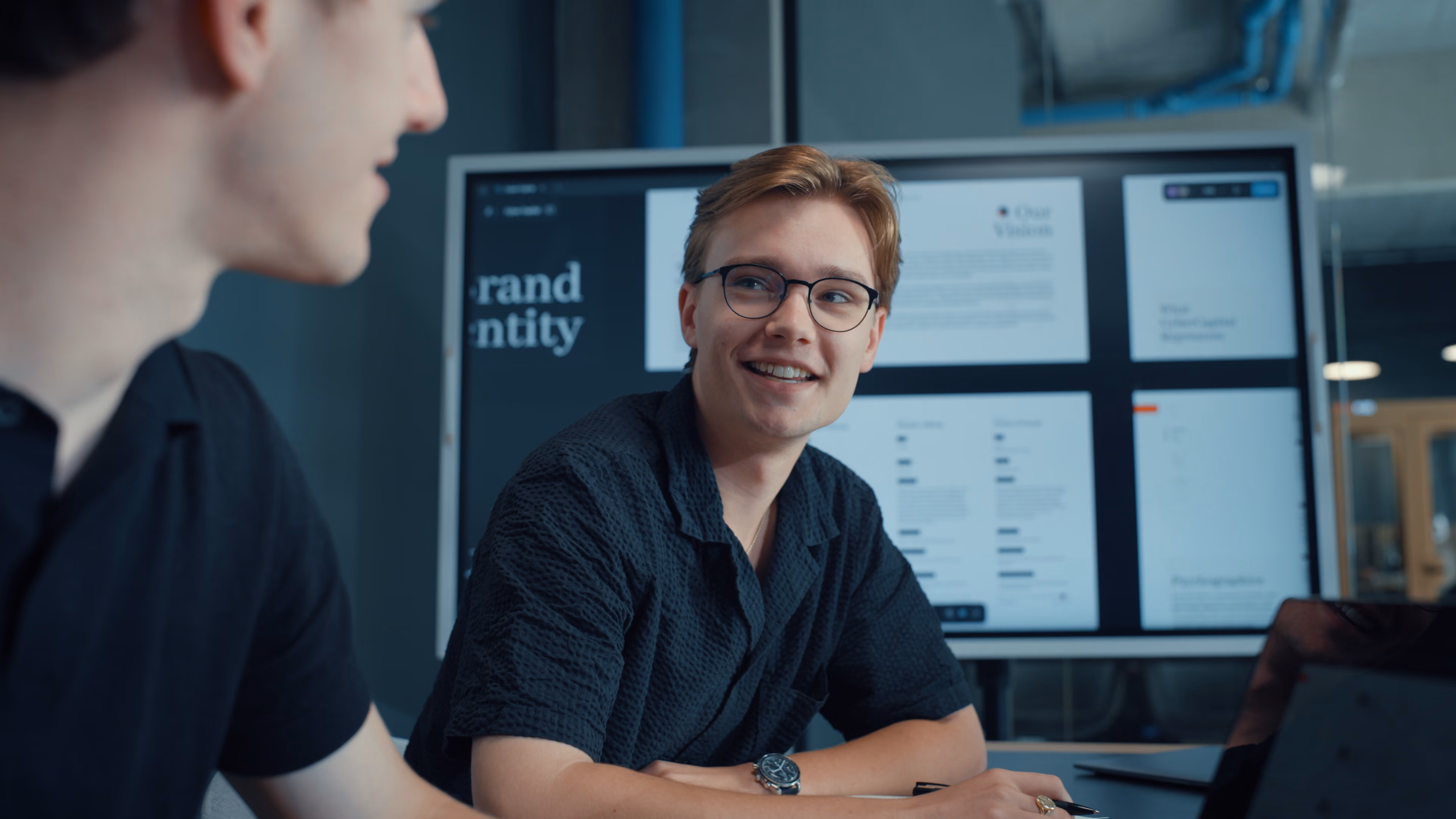 Twee jonge mannen bespreken merkidentiteitsplannen met een presentatie op een scherm in een kantoor.