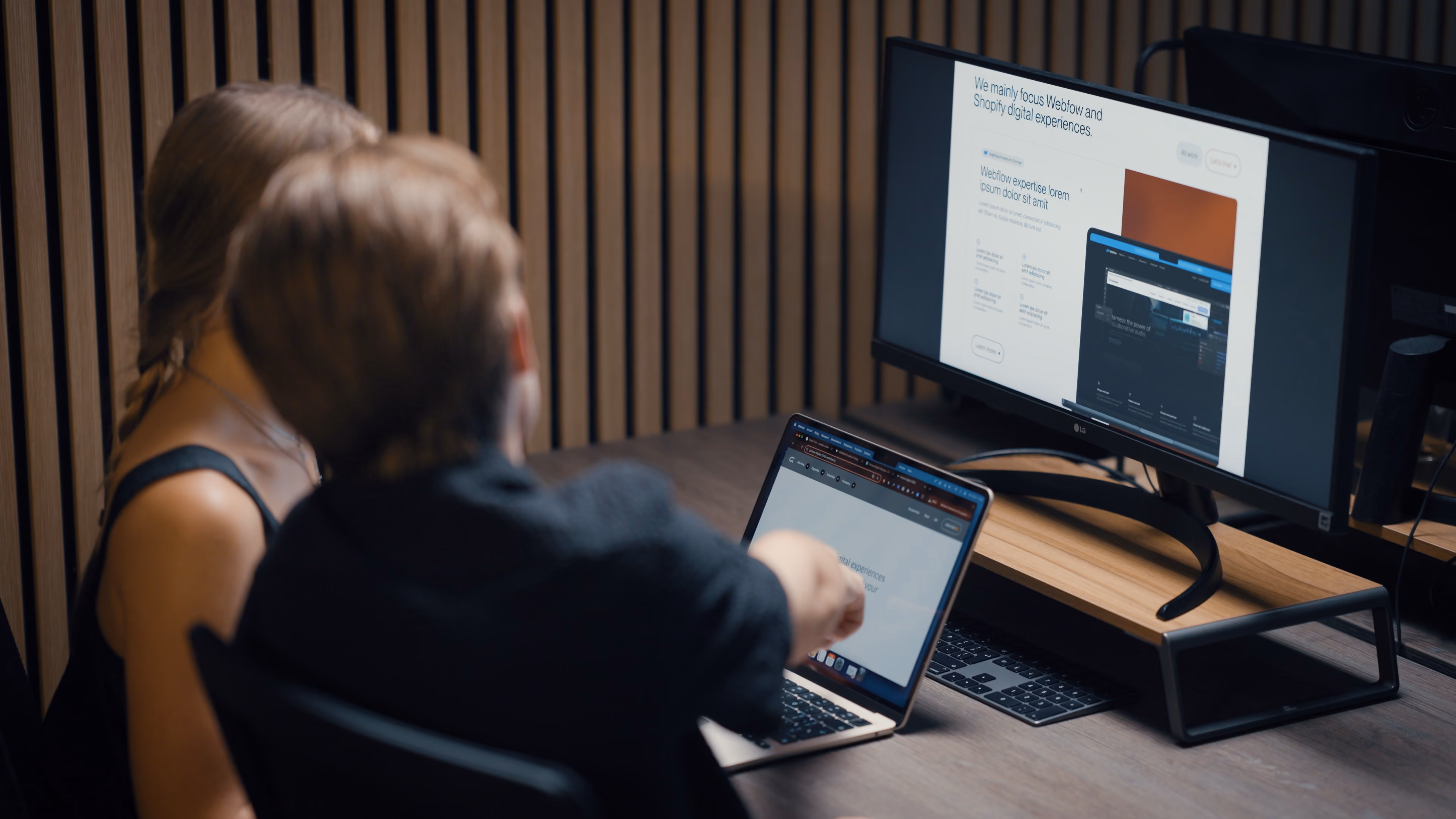 Twee mensen kijken naar een laptop en een grote monitor met website-ontwerp op een bureau.
