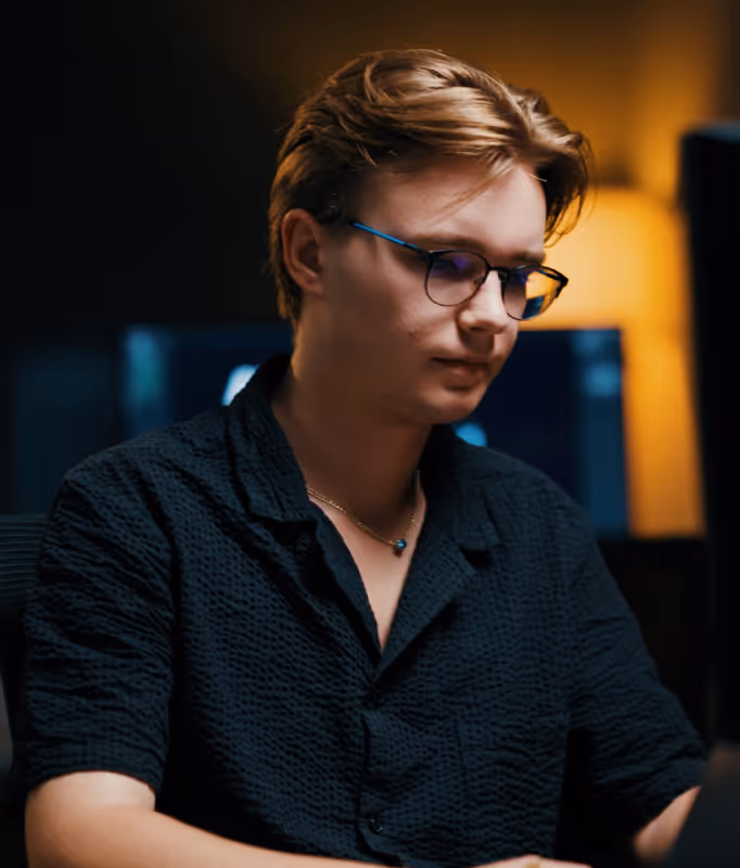 Jonge man met bril en zwart shirt werkt geconcentreerd in een schemerige kamer met warme verlichting.