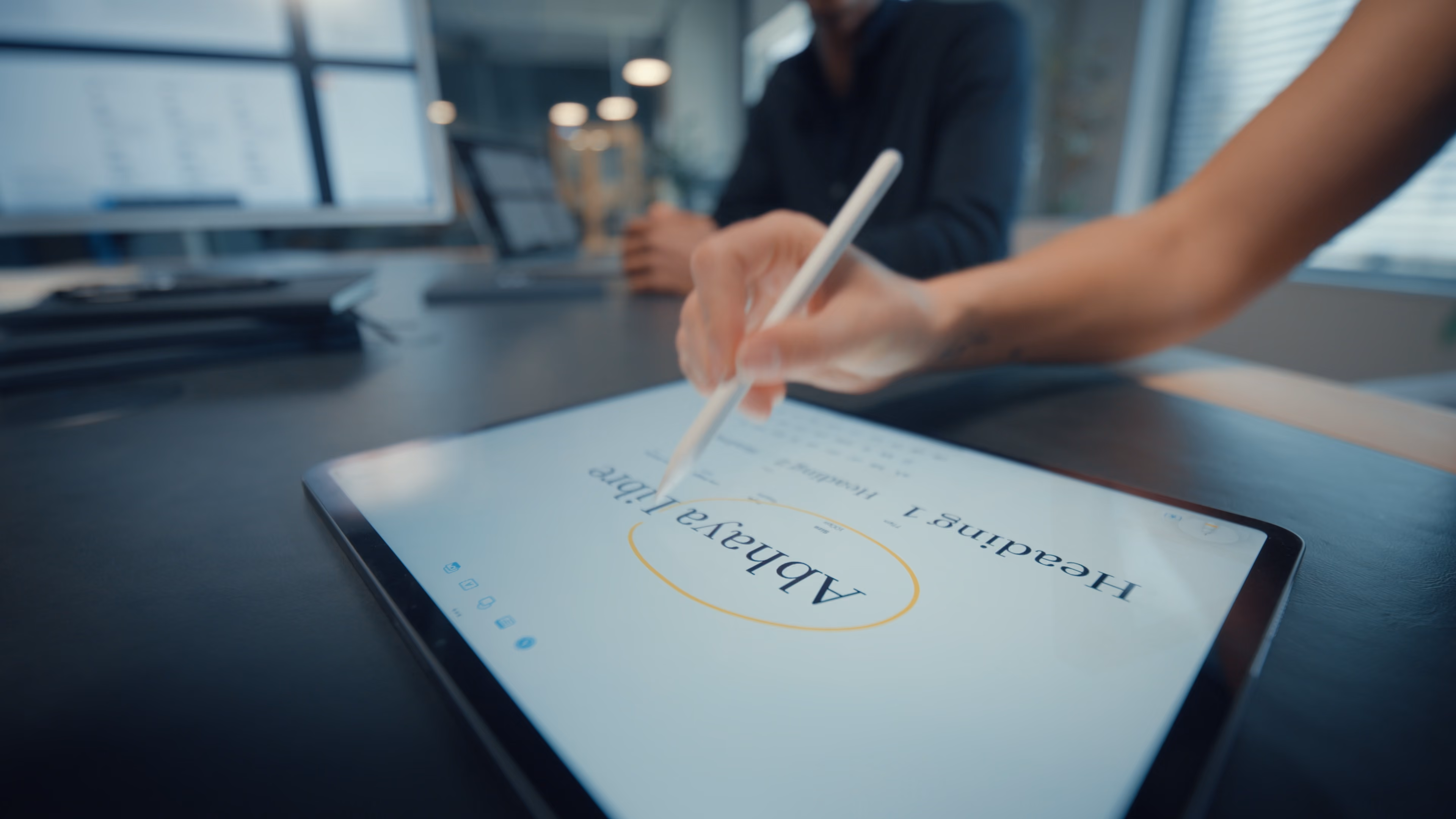Hand uses white stylus to circle text on a tablet screen in an office setting.