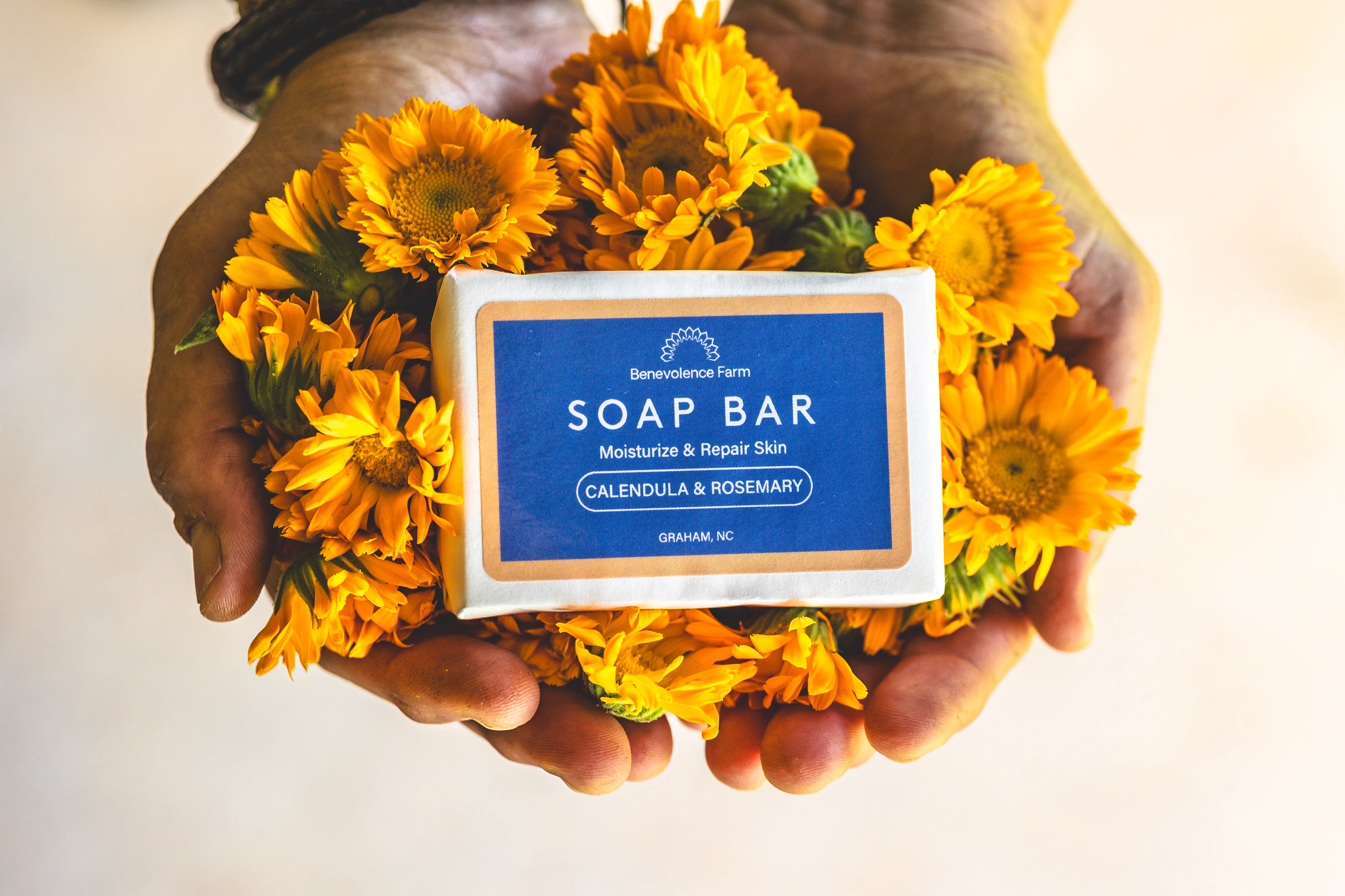Hands holding a farm-made soap bar surrounded by marigolds