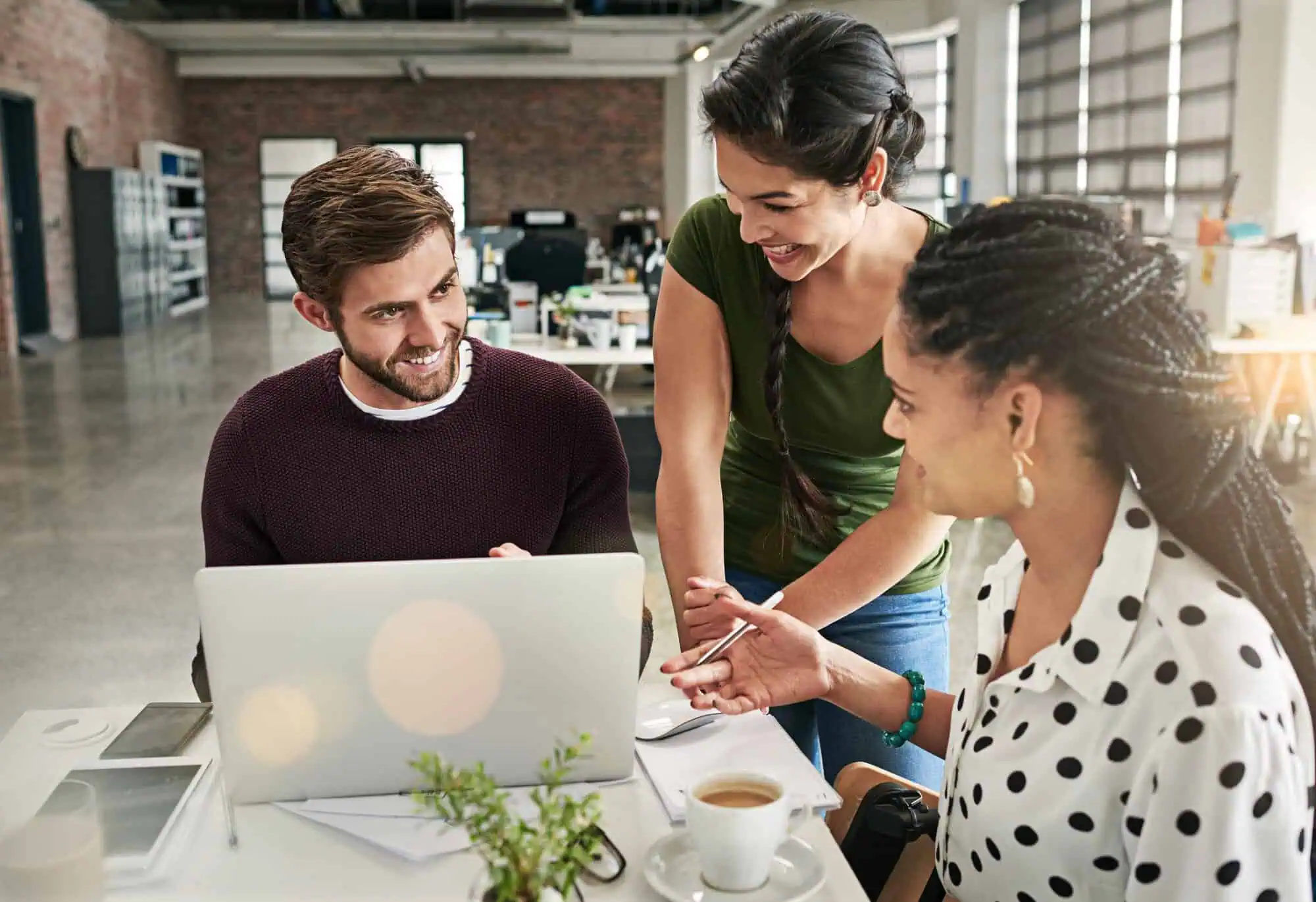 Three coworkers smiling and discussing work around a laptop in a modern office space.