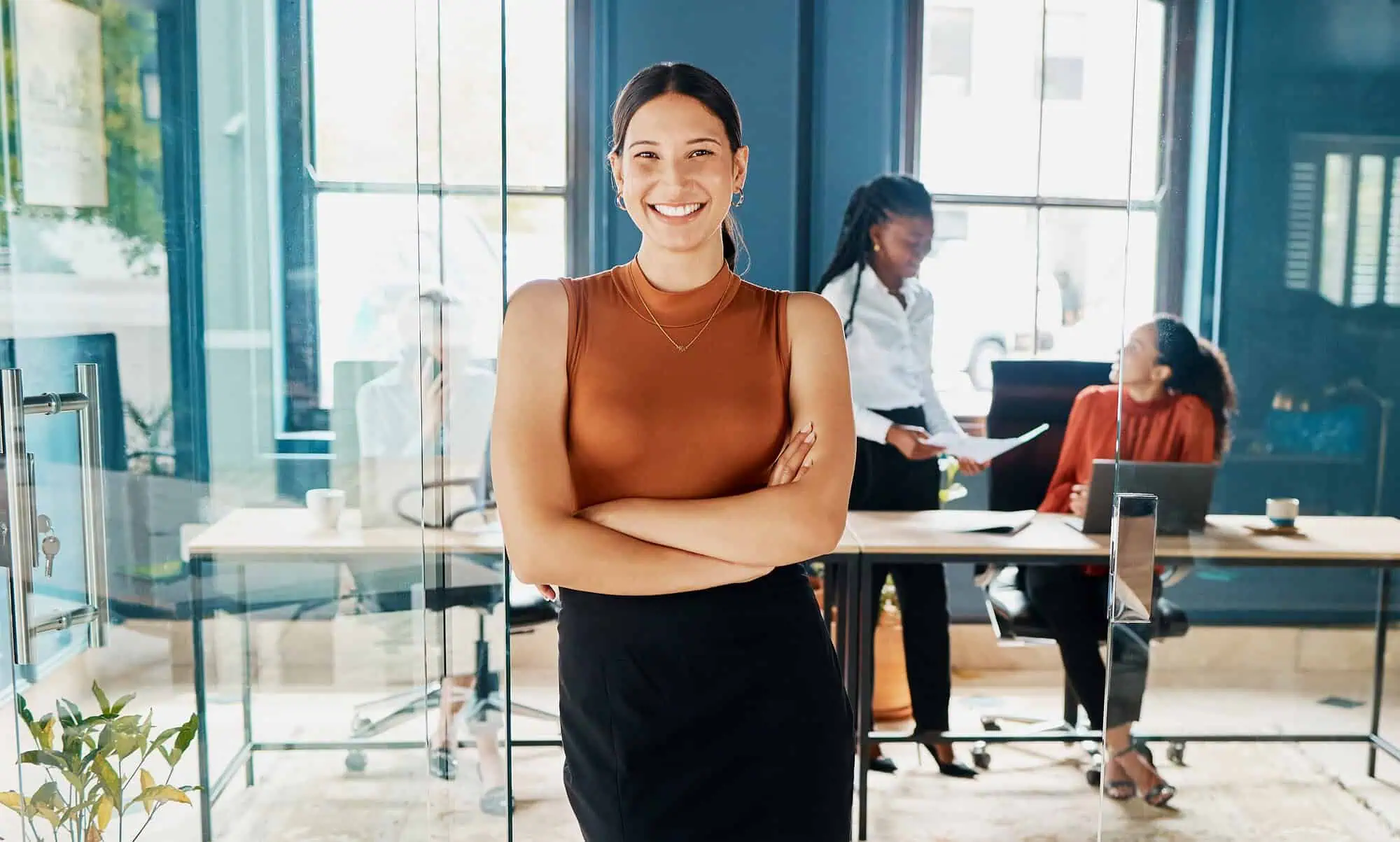 Smiling woman in brown top standing confidently with colleagues working behind in an office.