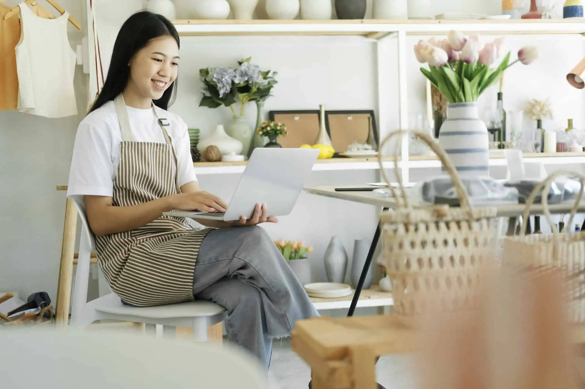 Smiling woman in apron sitting with crossed legs using laptop in a decorated home or shop space.