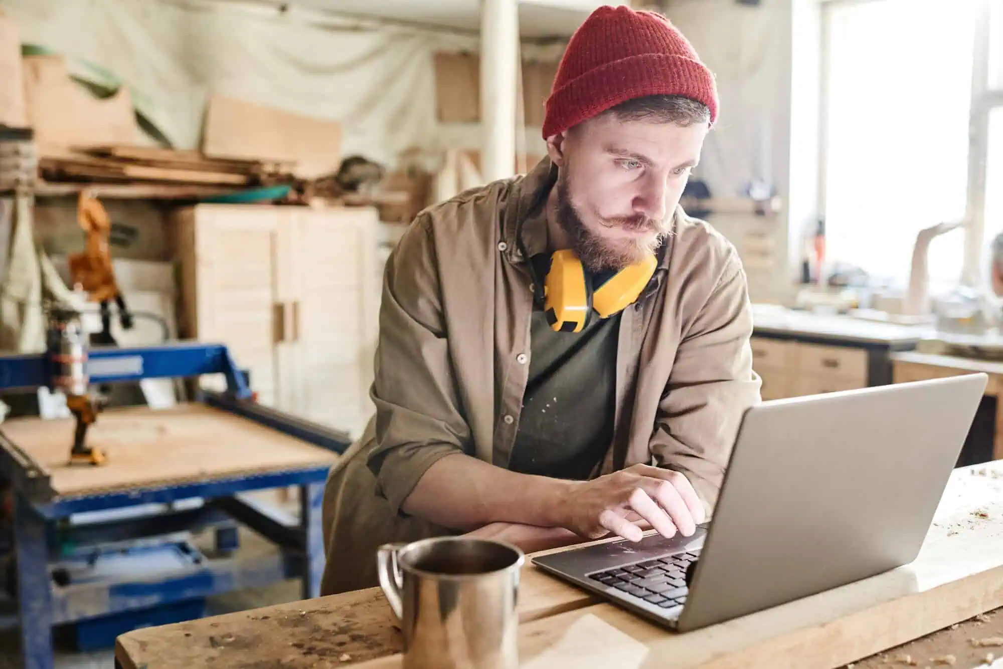 Man in a red beanie using a laptop in a woodworking shop with yellow ear protectors around his neck.