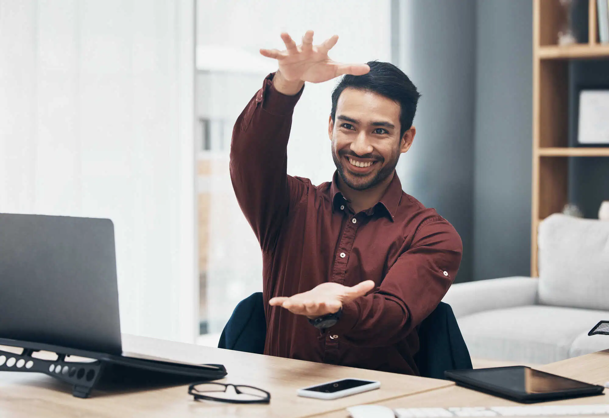 Smiling man in maroon shirt gestures with hands as if holding an invisible object at desk with laptop.