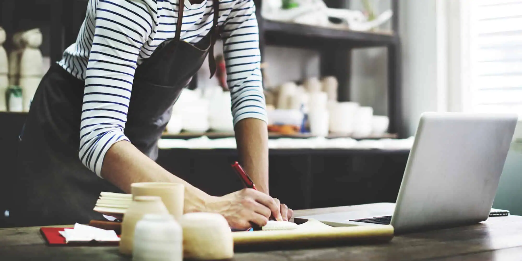 Person in striped shirt and apron writing on paper at table with pottery and laptop nearby.