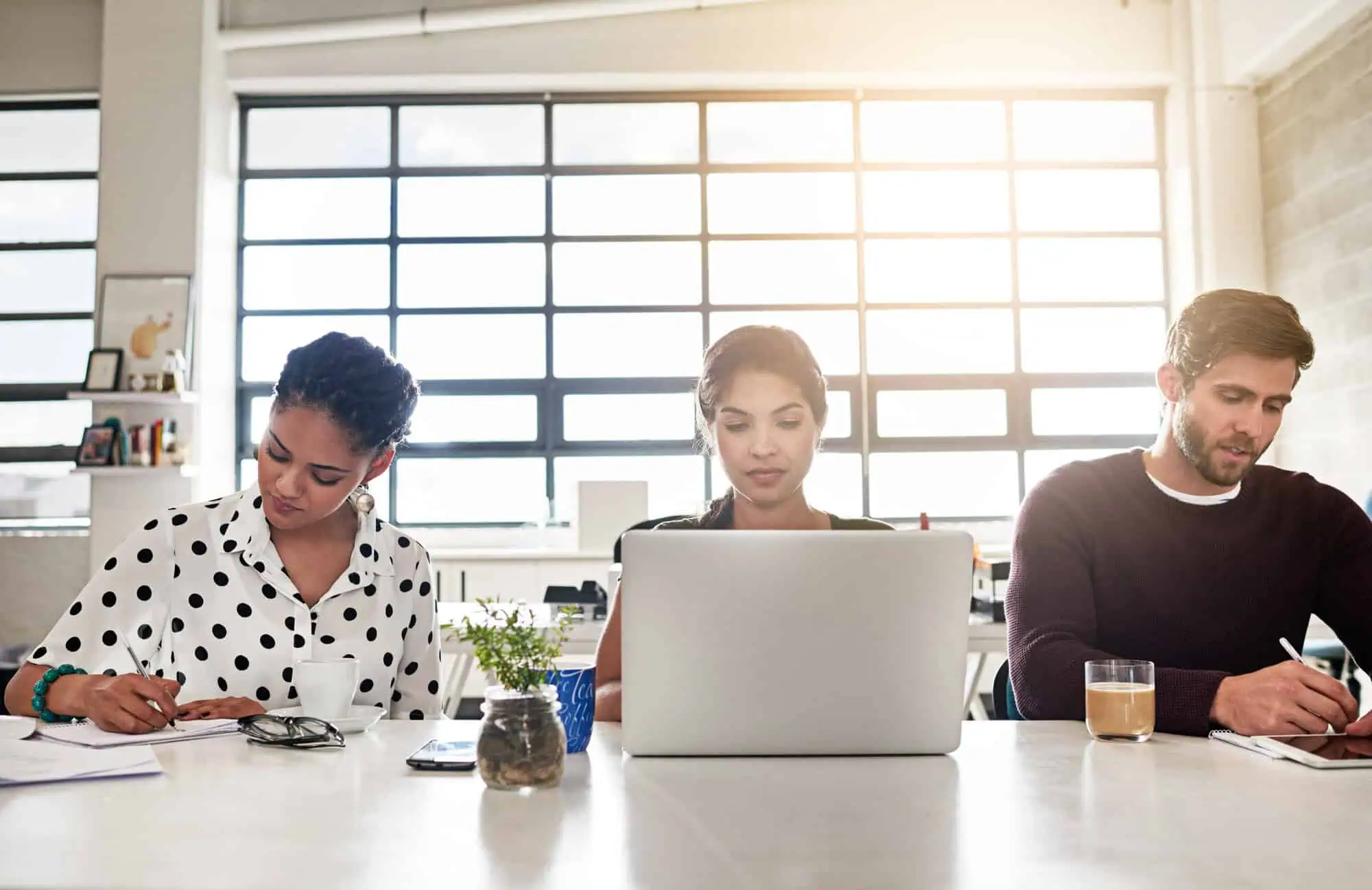 Three diverse coworkers focused on work at a bright office desk with laptop, notes, and drinks.