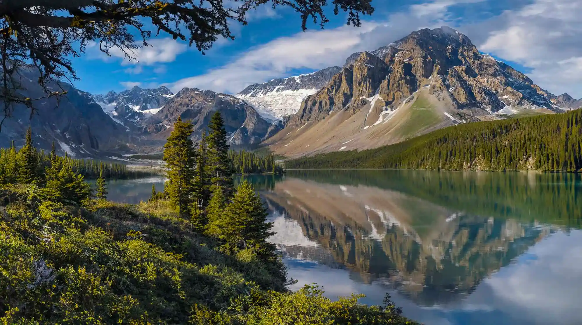 Mountain range with snow patches reflecting in a clear lake surrounded by green forest and blue sky.