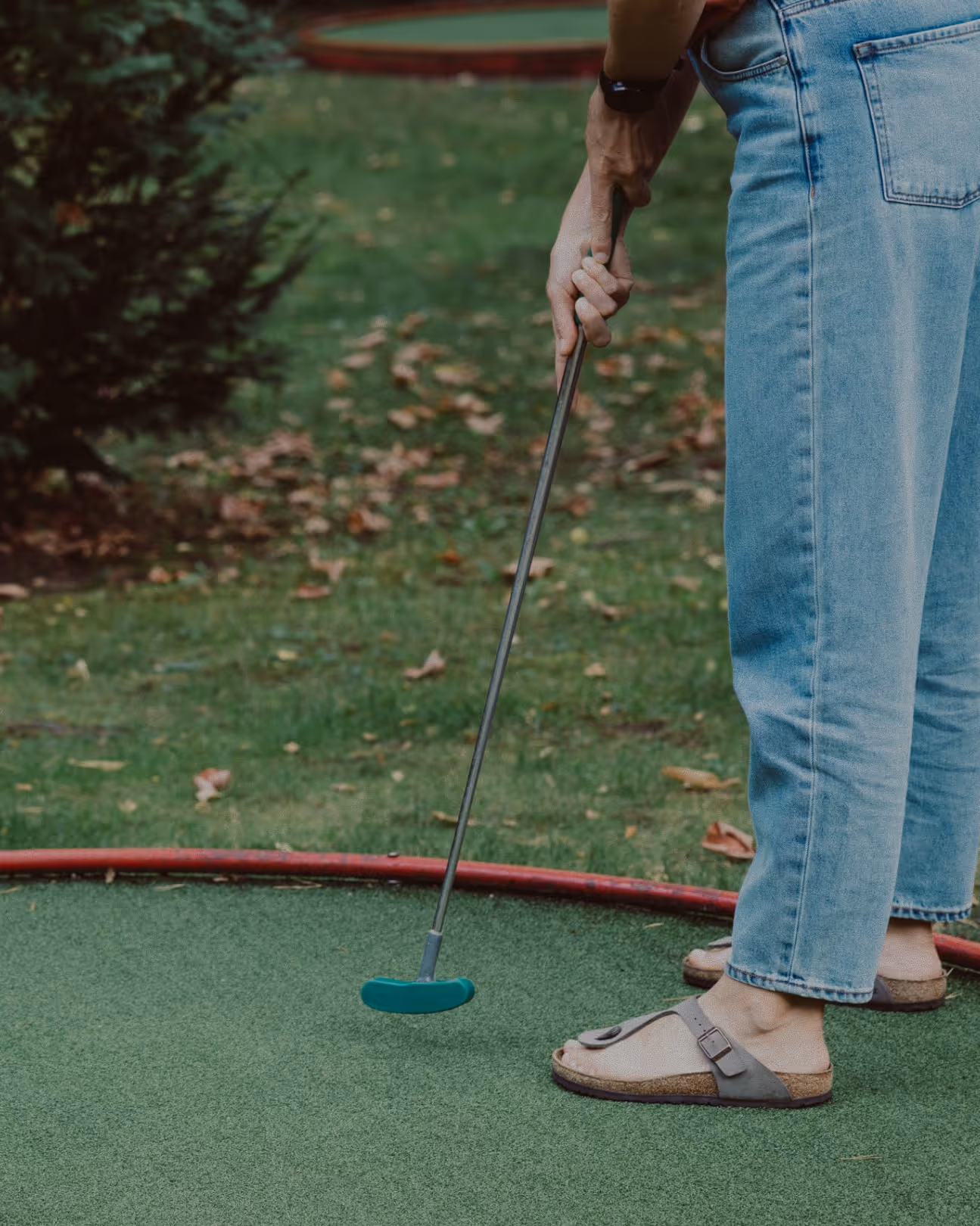 Person holding a blue mini golf putter standing on a green mini golf course surrounded by grass and bushes.