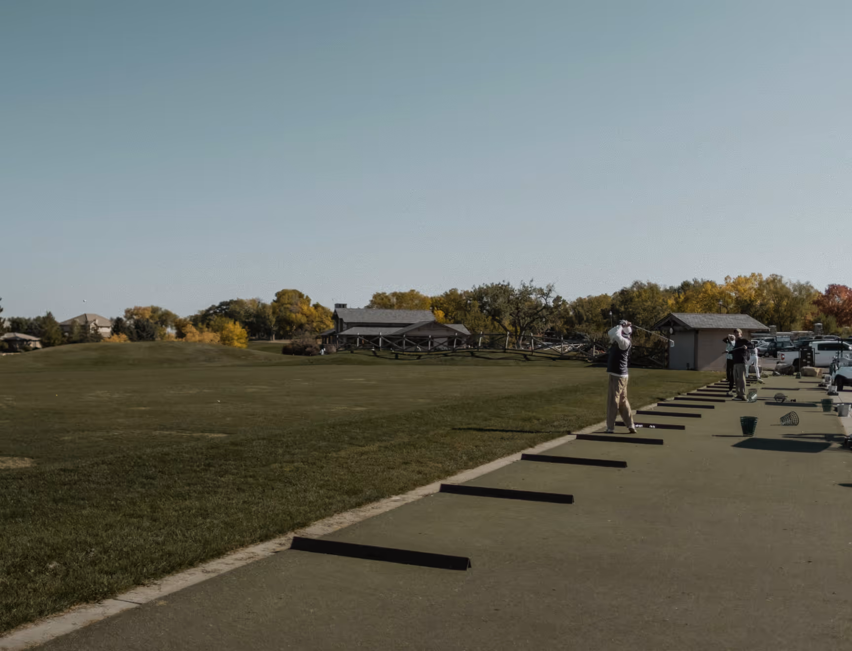 People practicing golf swings at an outdoor driving range with green grass and trees in the background.