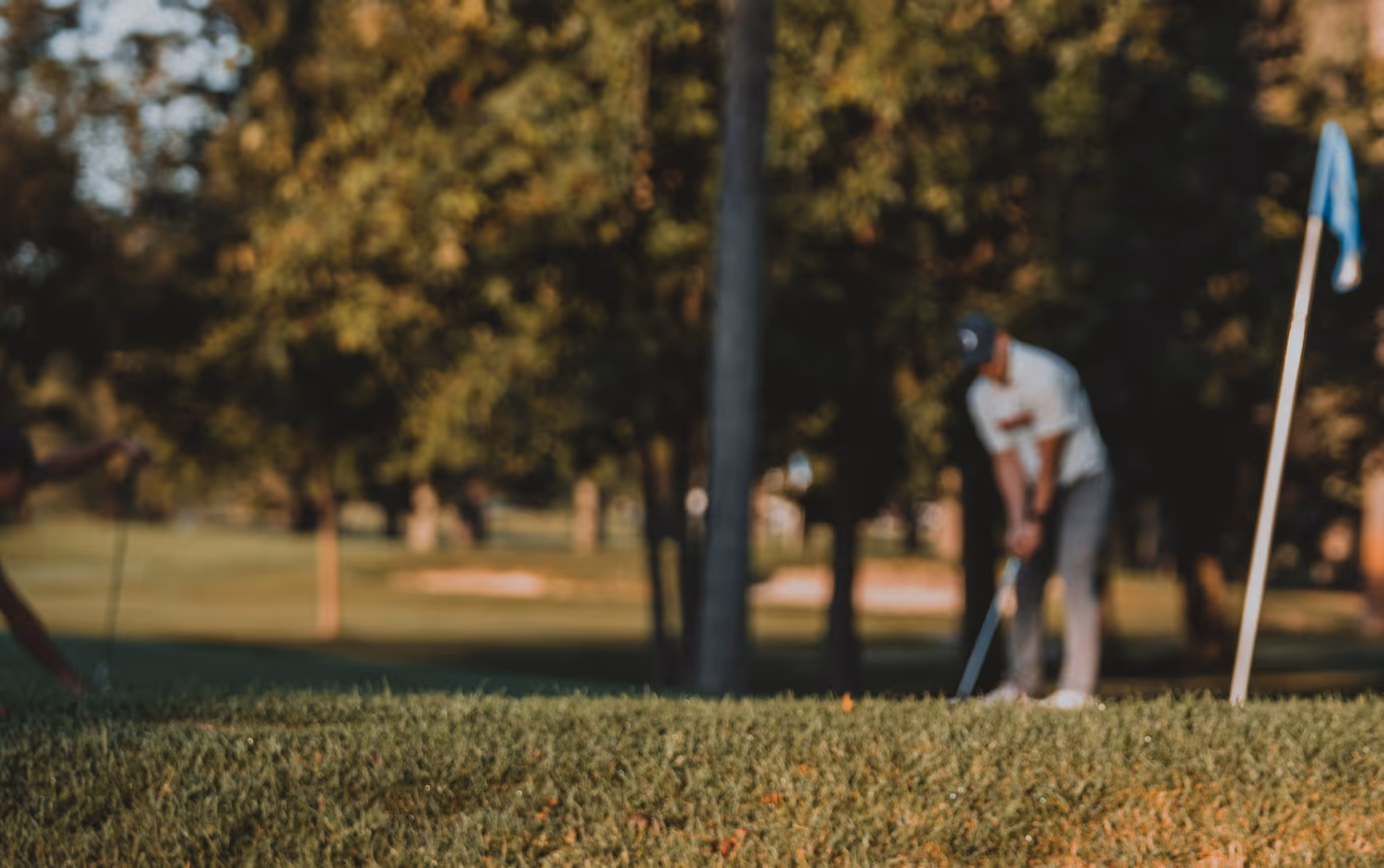 Golfer preparing to putt near a flagstick on a golf course with trees in the background, image is blurred.