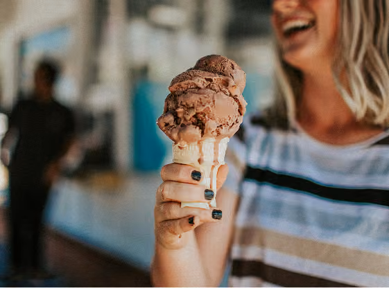 Close-up of a person holding a chocolate ice cream cone with two scoops, smiling in a casual striped shirt.