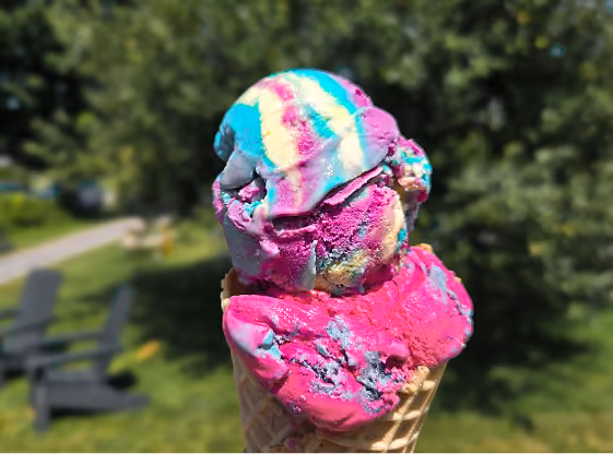 Close-up of a waffle cone holding two scoops of colorful ice cream in shades of pink, blue, yellow, and white, with a blurred green outdoor background.