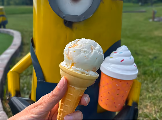 Hand holding a vanilla ice cream cone with orange streaks in front of a yellow Minion character statue holding a toy ice cream cone in an outdoor park.