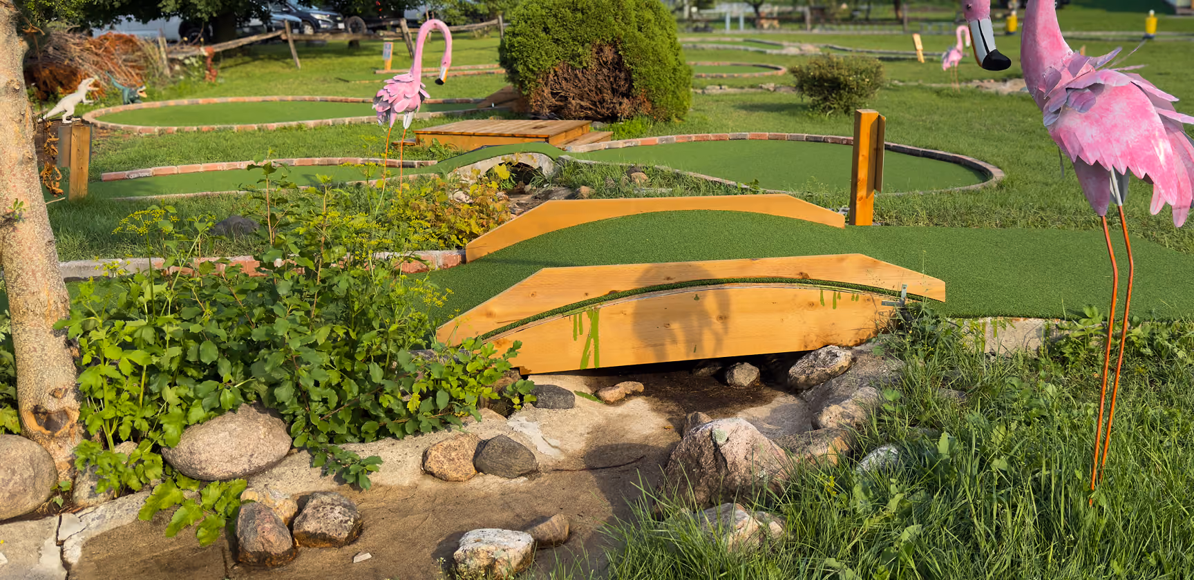 Outdoor mini golf course with green turf paths and curved wooden bridges over small rocky streams, decorated with pink flamingo statues and surrounded by grass and bushes.