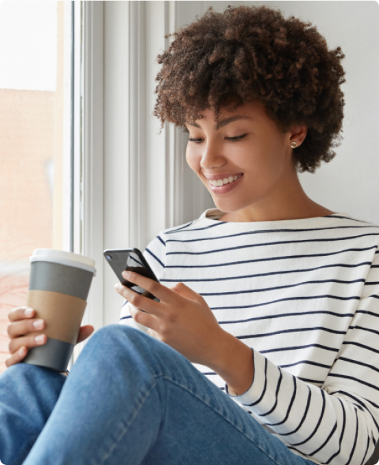 Smiling woman wearing striped shirt, holding a coffee cup and looking at her phone by a window.