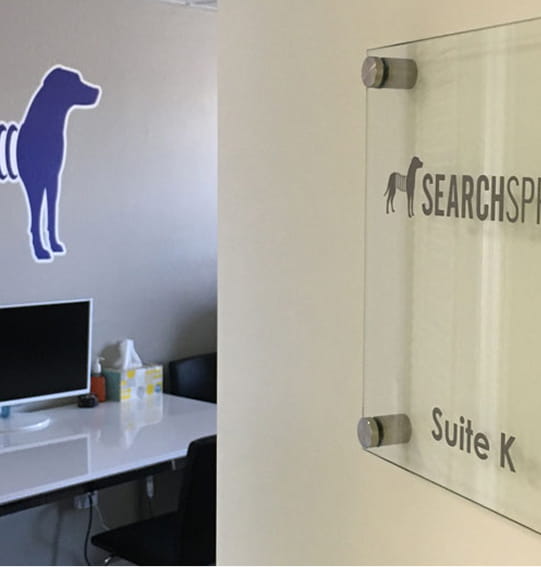 Office entrance with a glass sign reading SearchSP Suite K and a dog logo, with a desk and monitor inside.