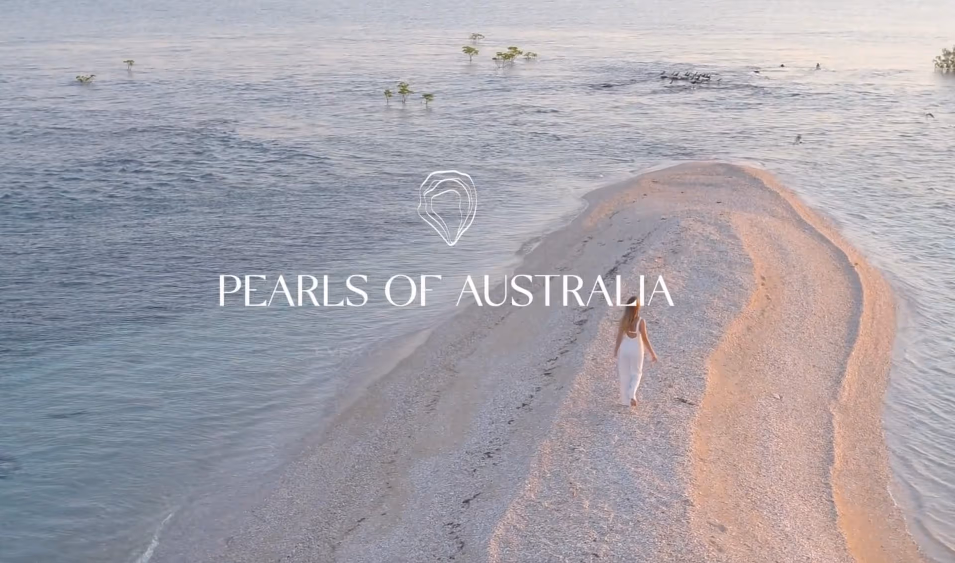 Woman in white walking alone on a sandy shoreline surrounded by calm water with small patches of vegetation.