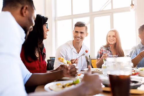 A diverse group of young adults sitting together at a table sharing a meal, smiling and engaging in conversation in a bright room with large windows.