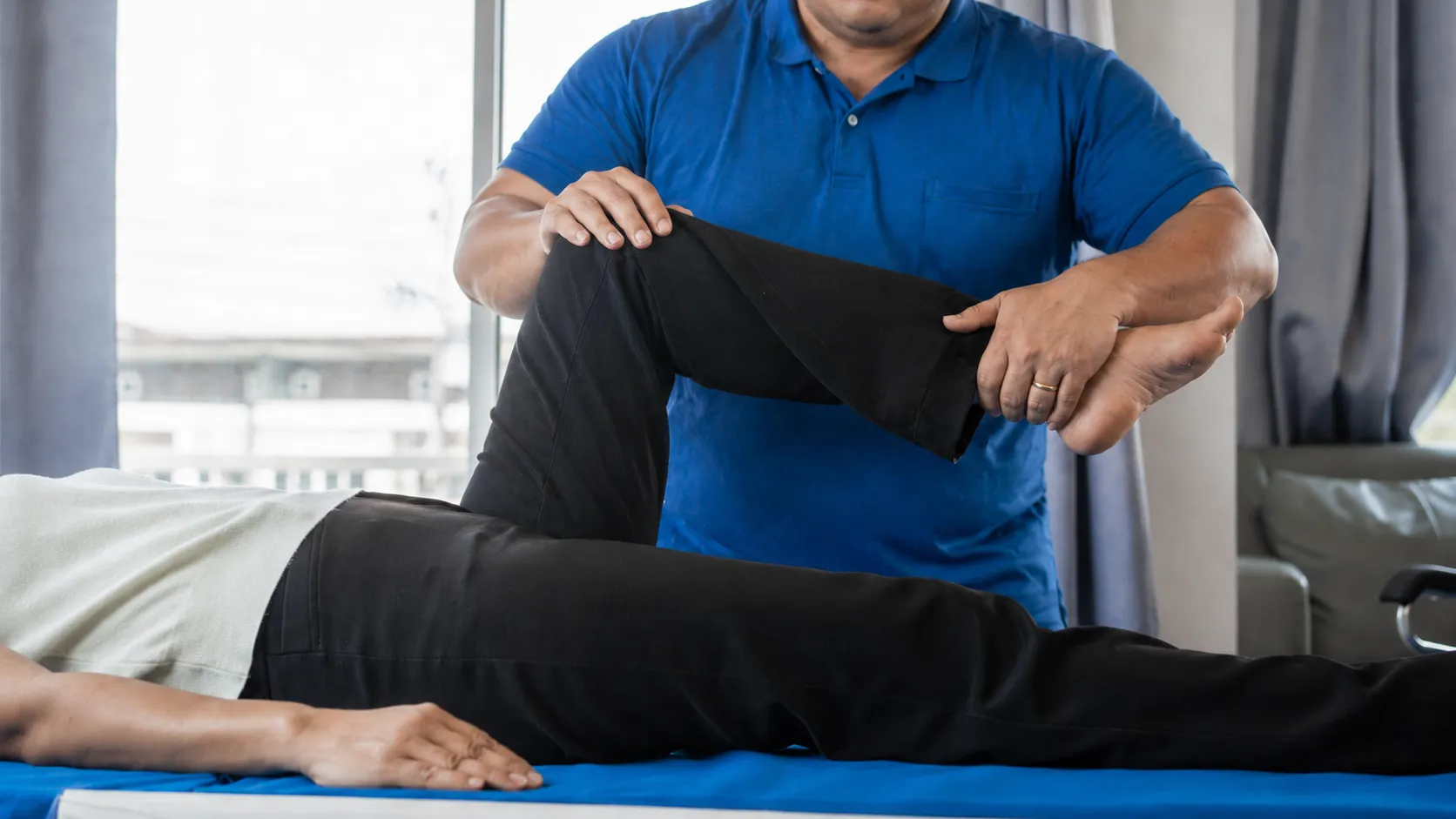 Therapist in blue shirt assists stretching a patient's bent leg on a therapy table.