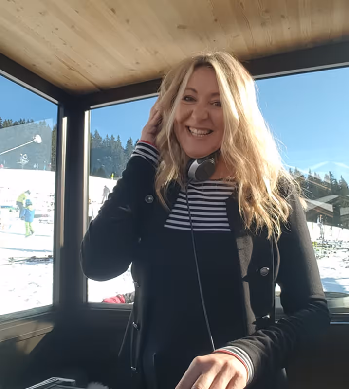 Lächelnde blonde Frau mit Kopfhörern um den Hals steht vor Fenstern mit verschneiter Landschaft und Skifahrern.