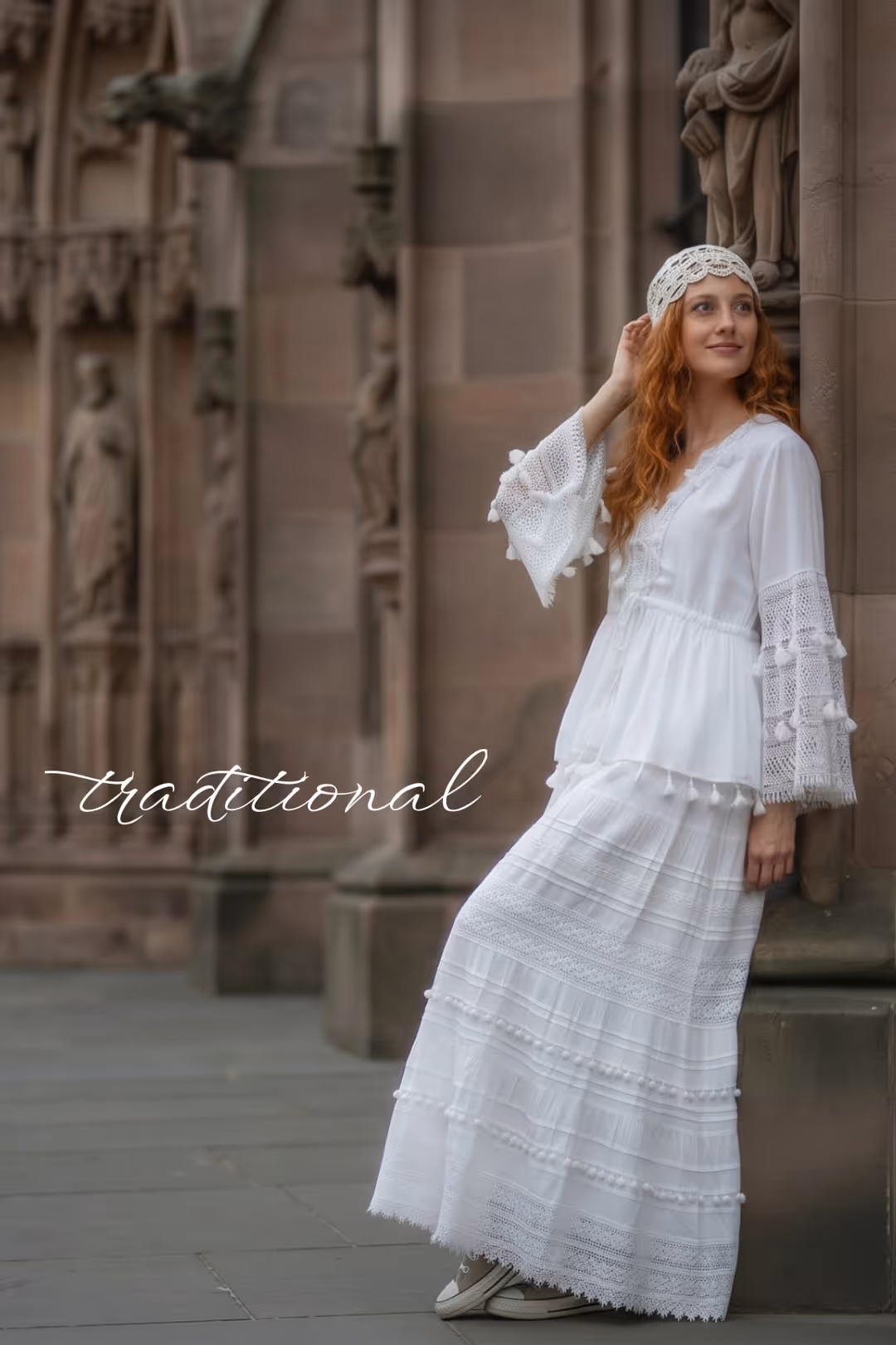 Frau in traditionellem weißem Kleid mit Häkelärmeln lehnt an Steinmauer neben historischer Fassade.