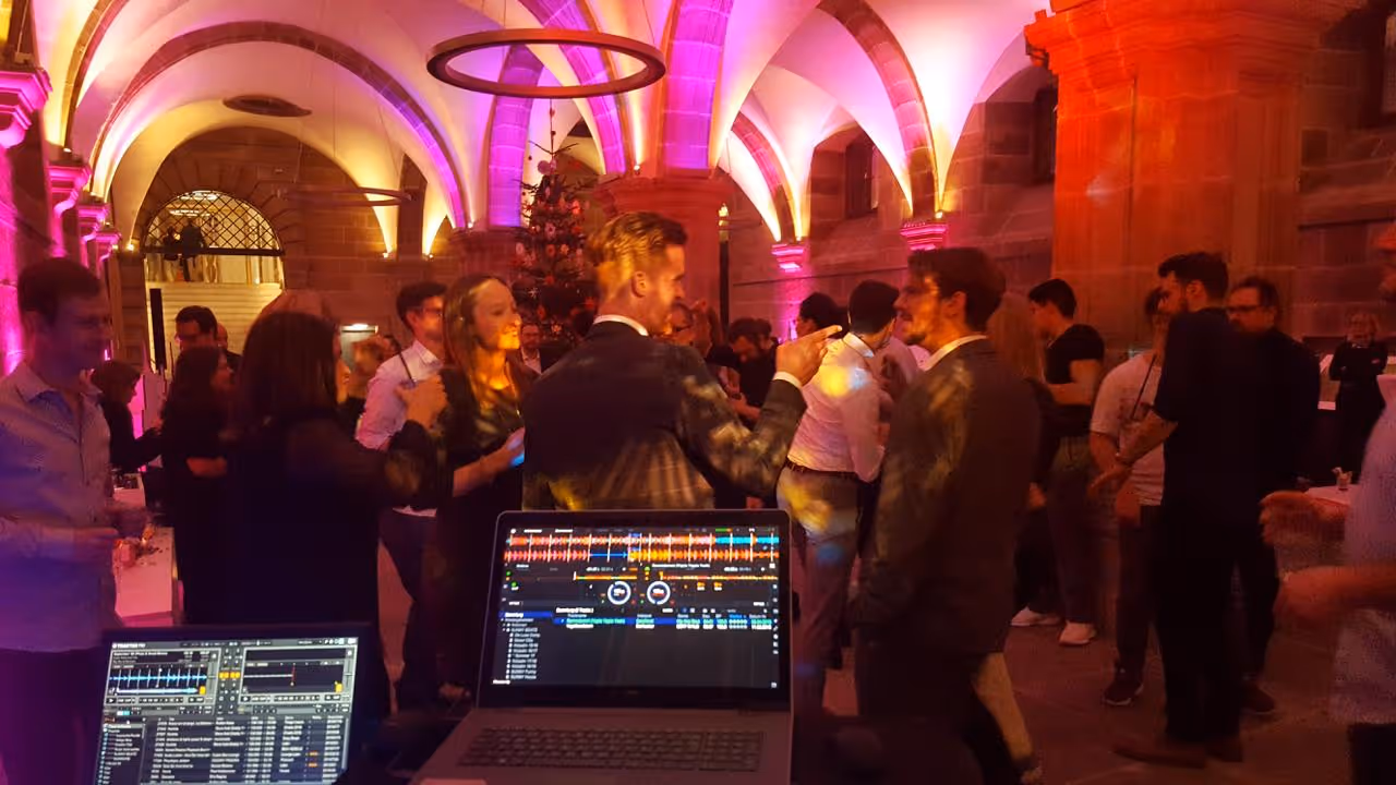 Menschen bei einer Party in einem festlich beleuchteten Raum mit Laptop-DJ-Setup im Vordergrund.
