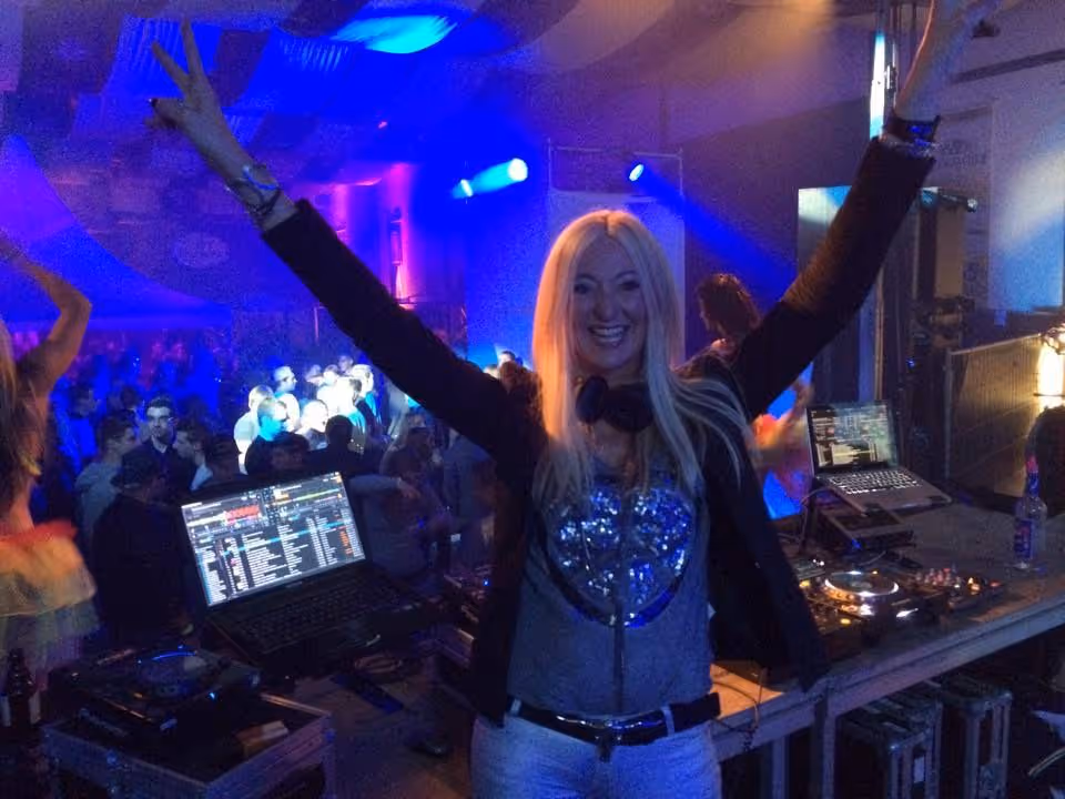DJ mit langem blondem Haar hebt die Arme in einer Clubumgebung mit blauer Beleuchtung.