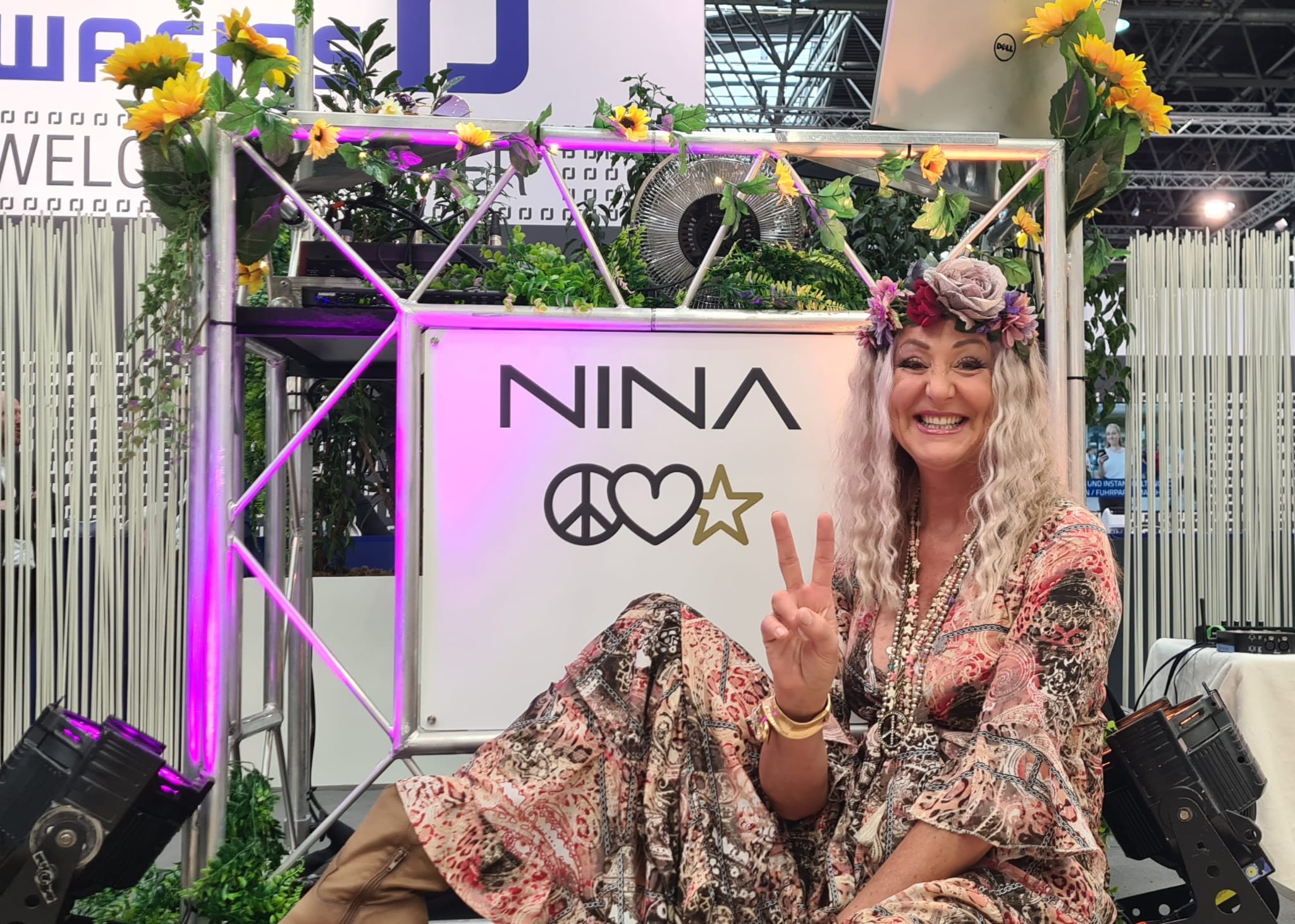 Fröhliche Frau mit Blumenschmuck im Haar zeigt Friedenszeichen vor einem dekorierten Stand mit der Aufschrift NINA.