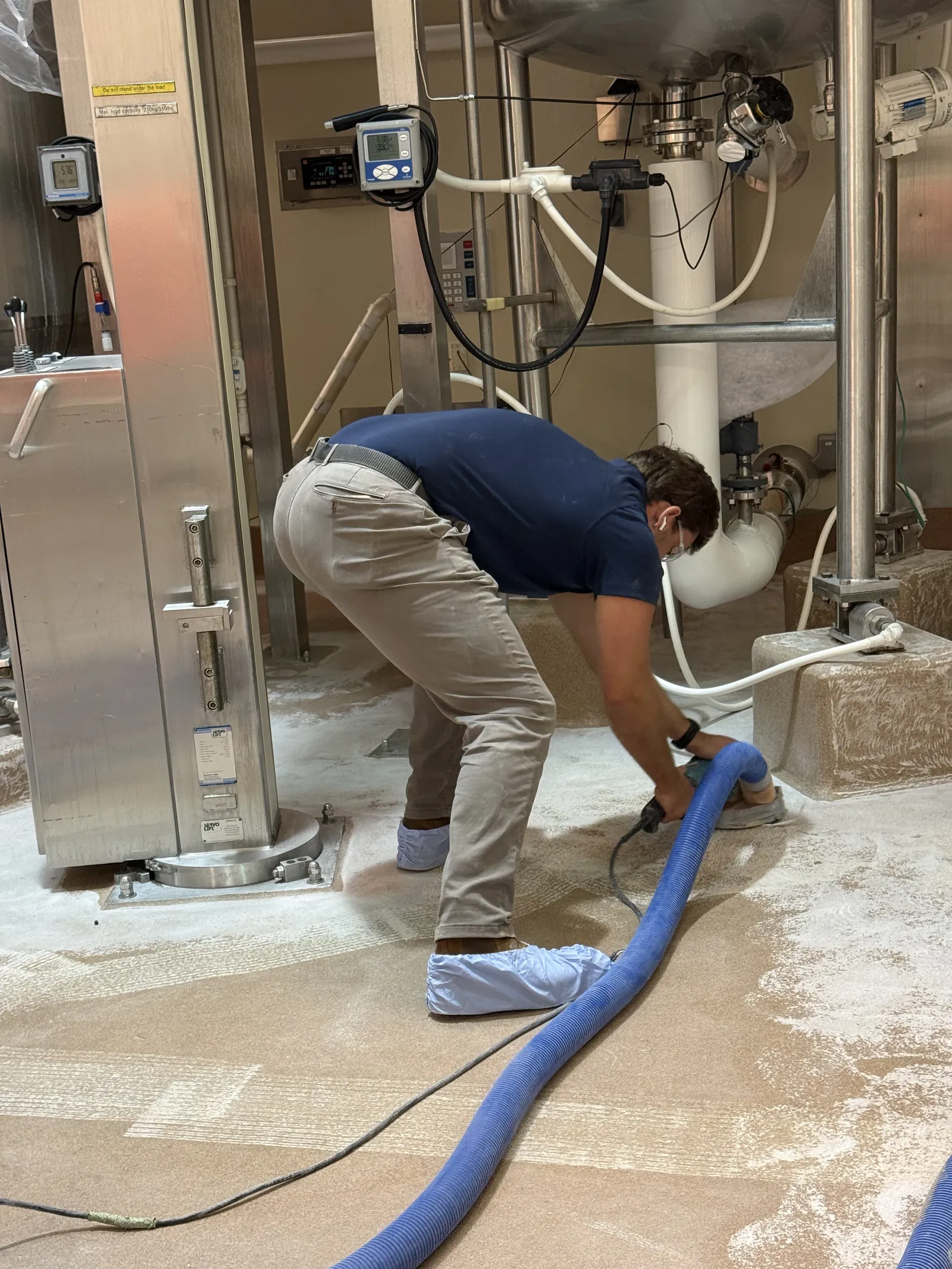 Installer running a dust-extraction vacuum at a stainless tank leg during floor prep