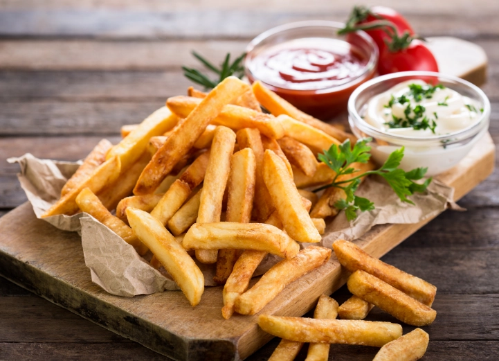 Golden French fries served on a wooden board with ketchup and mayonnaise in small glass bowls, garnished with parsley and cherry tomatoes.