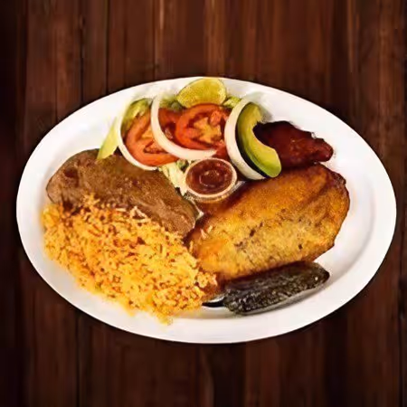 Plate with breaded fish, yellow rice, grilled meat, salad with tomato, onion, avocado, lime, a small cup of sauce, and a grilled pepper on a wooden table.