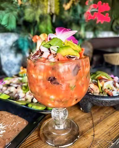 Glass of octopus cocktail garnished with avocado slices and an edible flower on a wooden table with a blurred background.