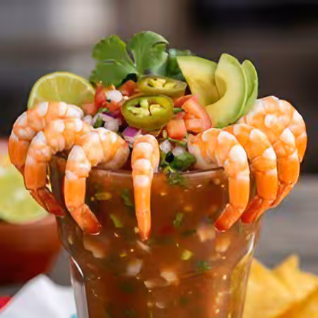 Glass of shrimp cocktail garnished with whole cooked shrimp, avocado slices, jalapeño, tomato salsa, lime wedge, and cilantro.