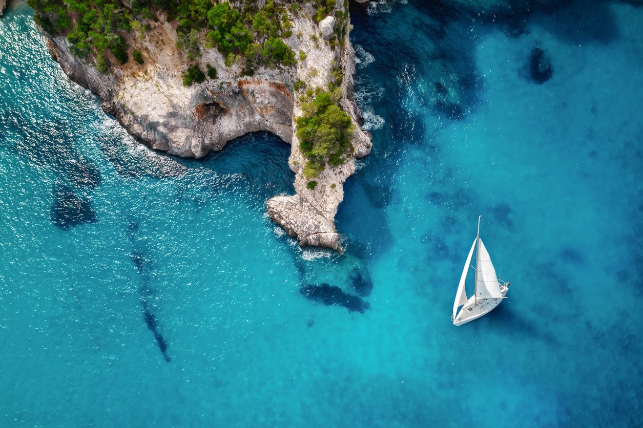 Aerial view of a sailboat anchored in turquoise water beside a rocky headland