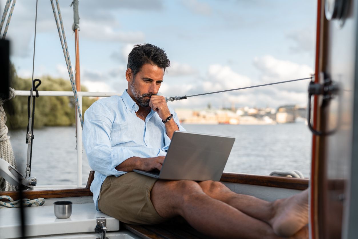 Man working on a laptop aboard a sailboat, looking thoughtful