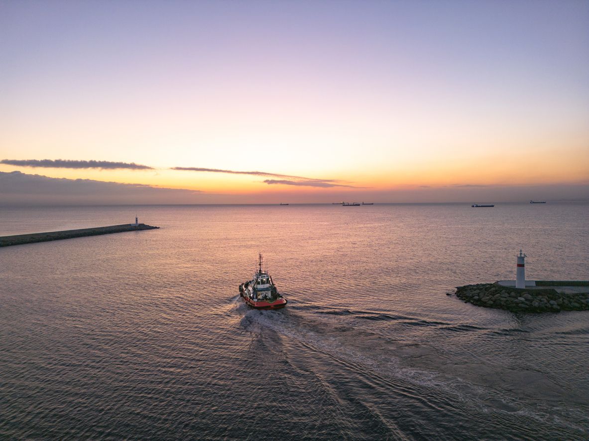 Tugboat heading out of a harbor at sunset past a small lighthouse