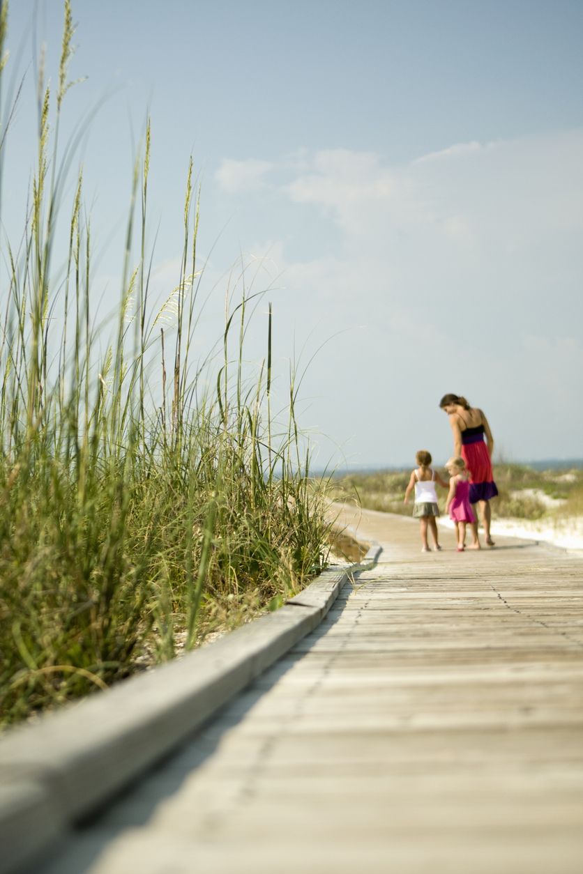 Adult and child walking along a wooden boardwalk through dune grass toward the beach