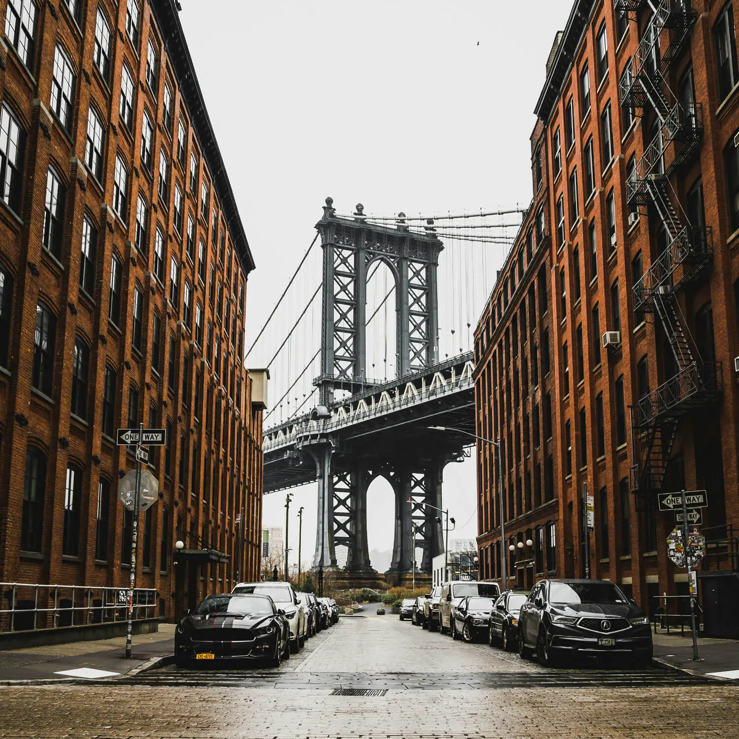 Manhattan Bridge framed by brick buildings and parked cars on street