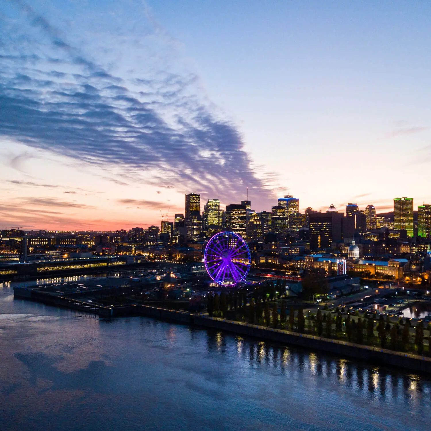 Illuminated Ferris wheel glows purple amid city skyline at twilight over river