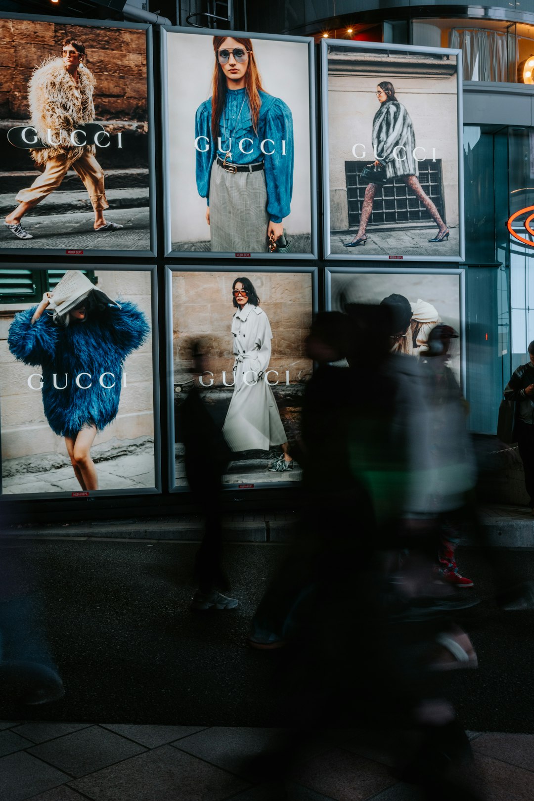 Pedestrians walking past a luxury fashion brand storefront with illuminated advertising displays at night in a shopping district
