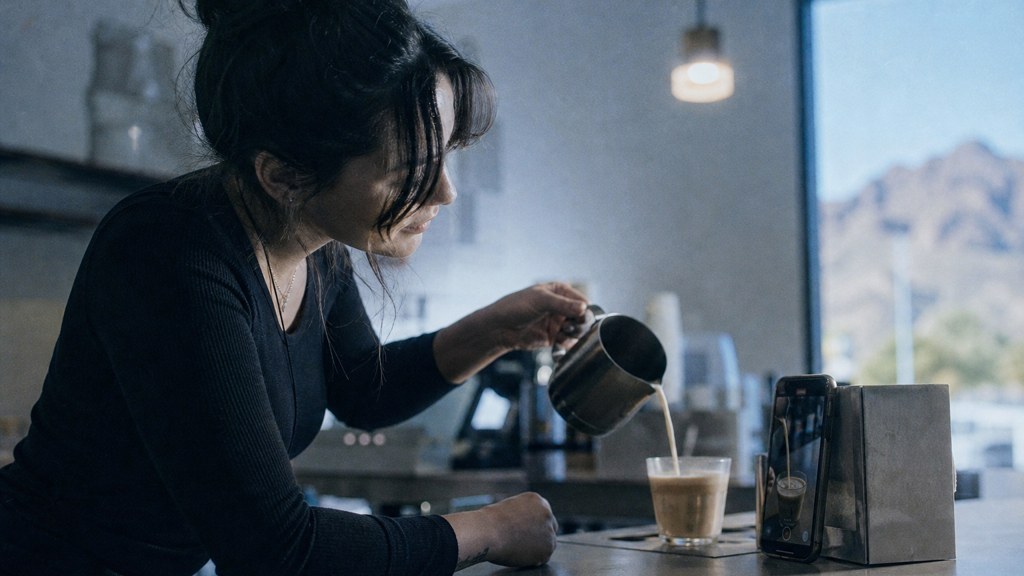 A young cafe worker filming a drink being poured on a phone propped against a napkin holder, photographed in Scottsdale