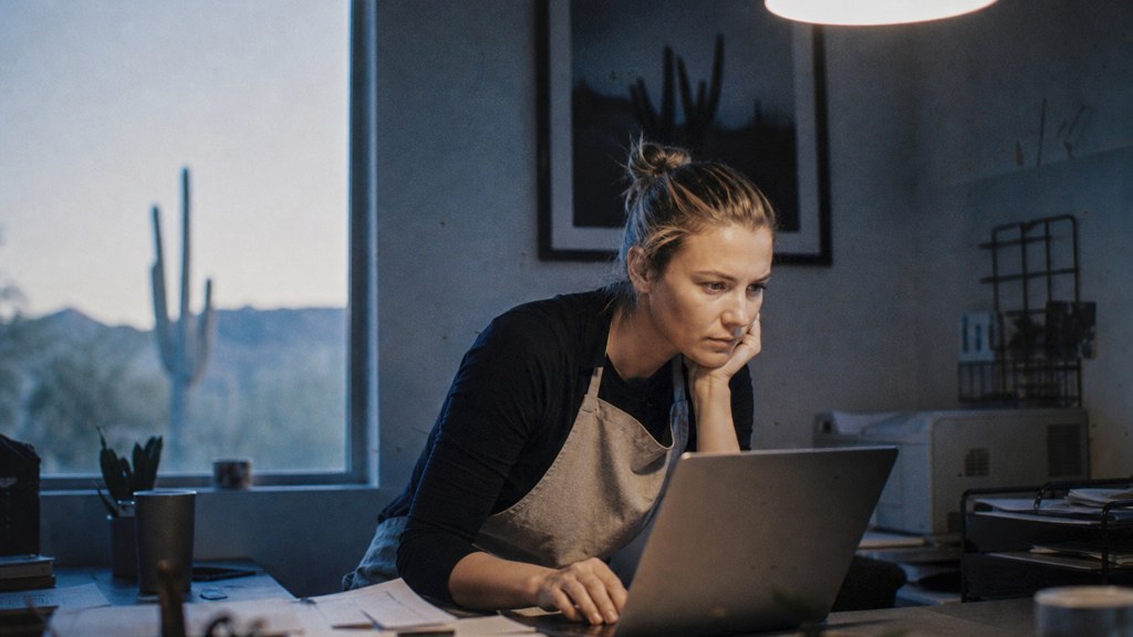 A small-business owner in an apron leaning over a laptop on a cluttered back-office desk late in the day, photographed in Scottsdale