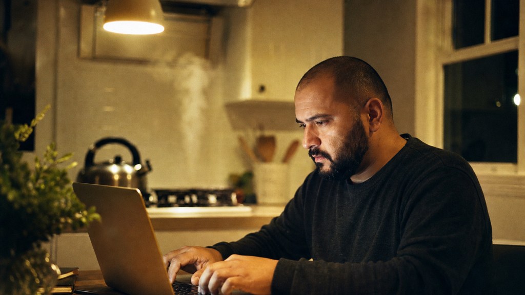 A service-business owner at a kitchen table typing a question into a laptop while a kettle steams behind, photographed in Santa Clarita