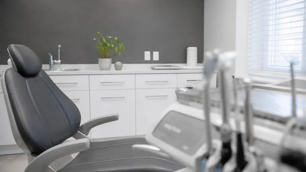 Editorial photograph of a tidy dental practice consultation room in neutral grey and white with cool daylight through blinds