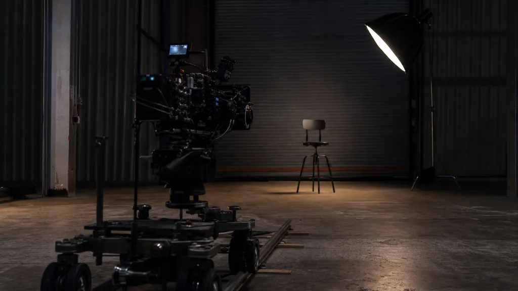 Editorial photograph of a warehouse film studio at dusk with a cinema camera on a dolly track facing an empty stool, one softbox and a roller door in deep shadow