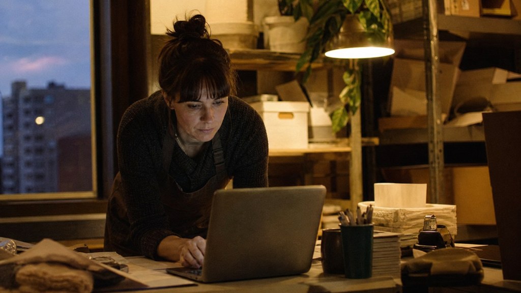 A small-business owner in an apron leaning over a laptop on a cluttered back-office desk late in the day, photographed in Spokane